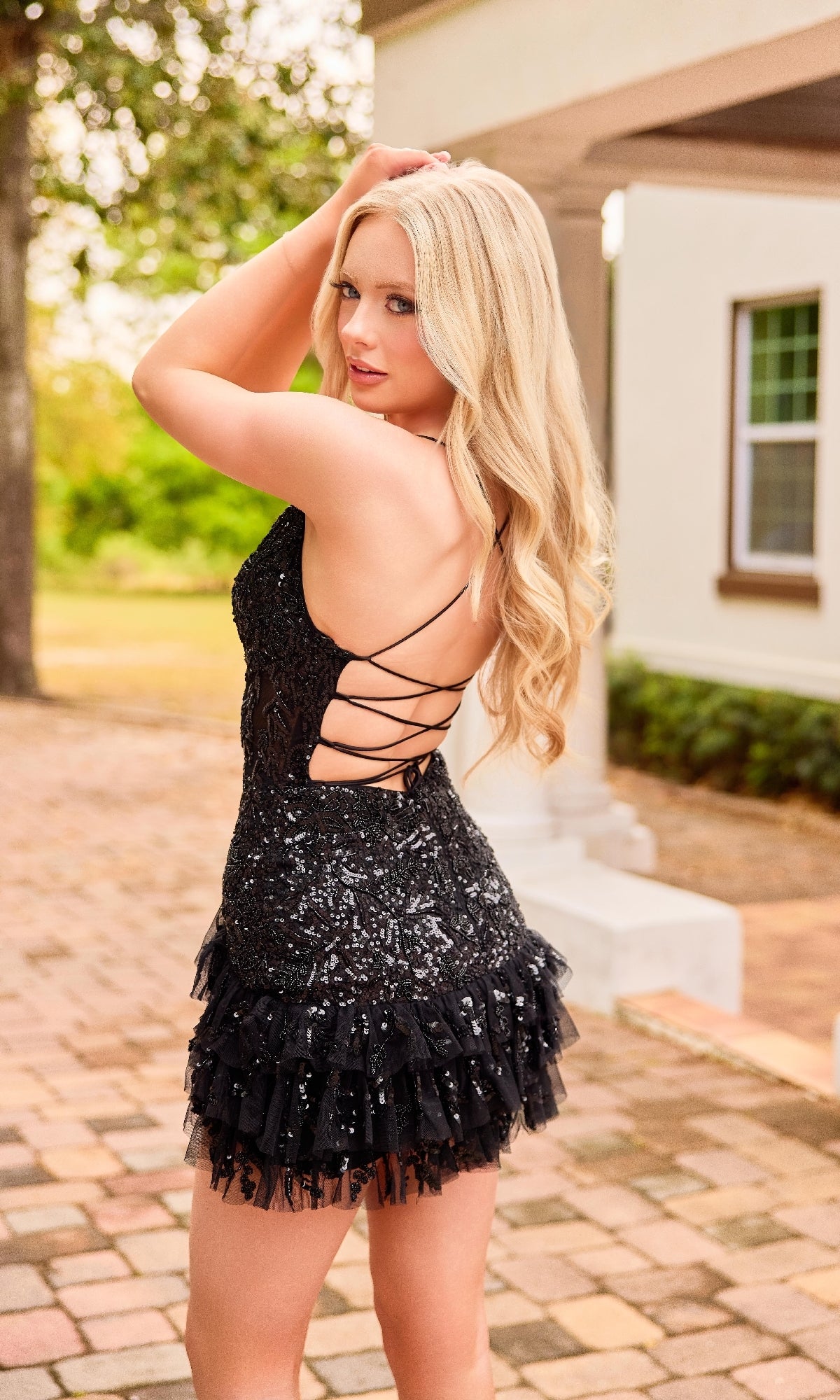 A woman with long blonde hair poses outdoors in the Primavera 4401, a short black sequin homecoming dress featuring a lace-up back and ruffled skirt—an ideal fit and flare style for special occasions.